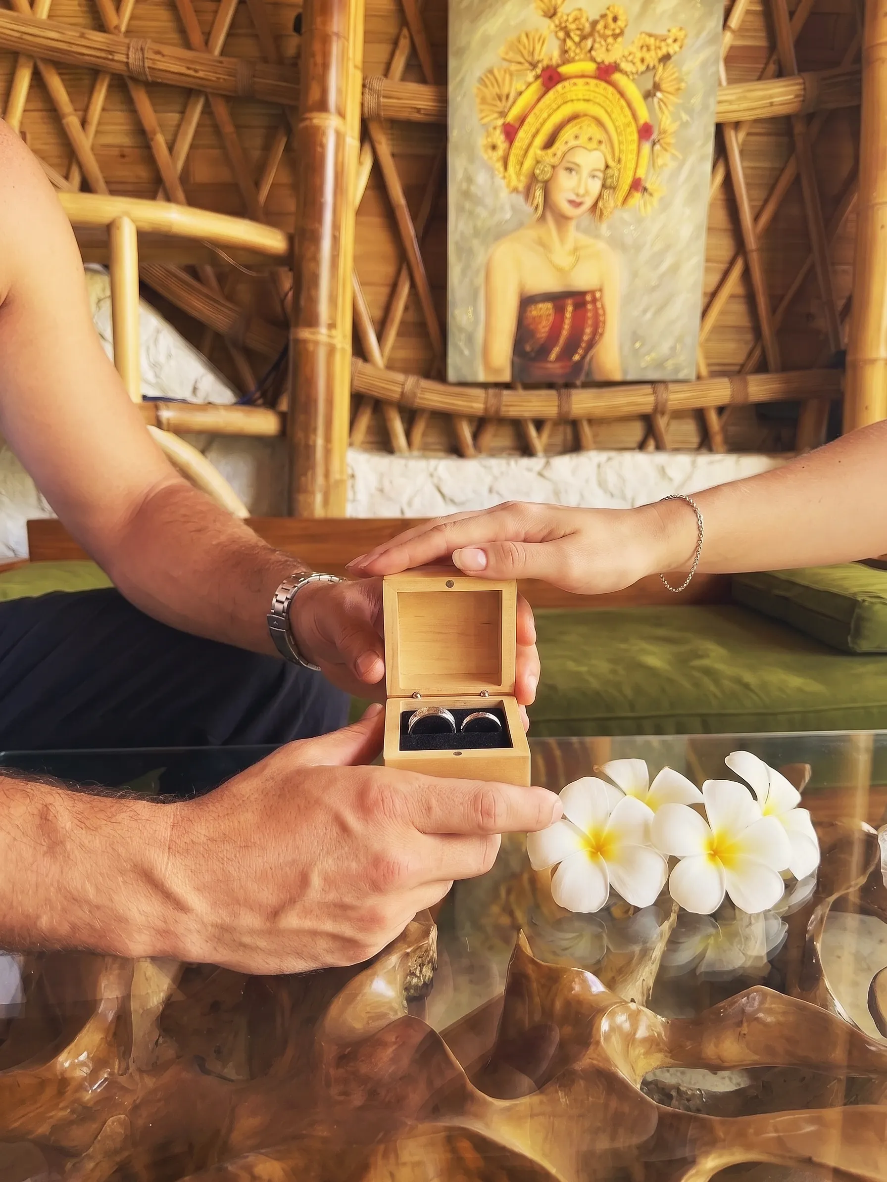 A couple open a wooden box with two matching hammered silver rings, frangipani on the glass table