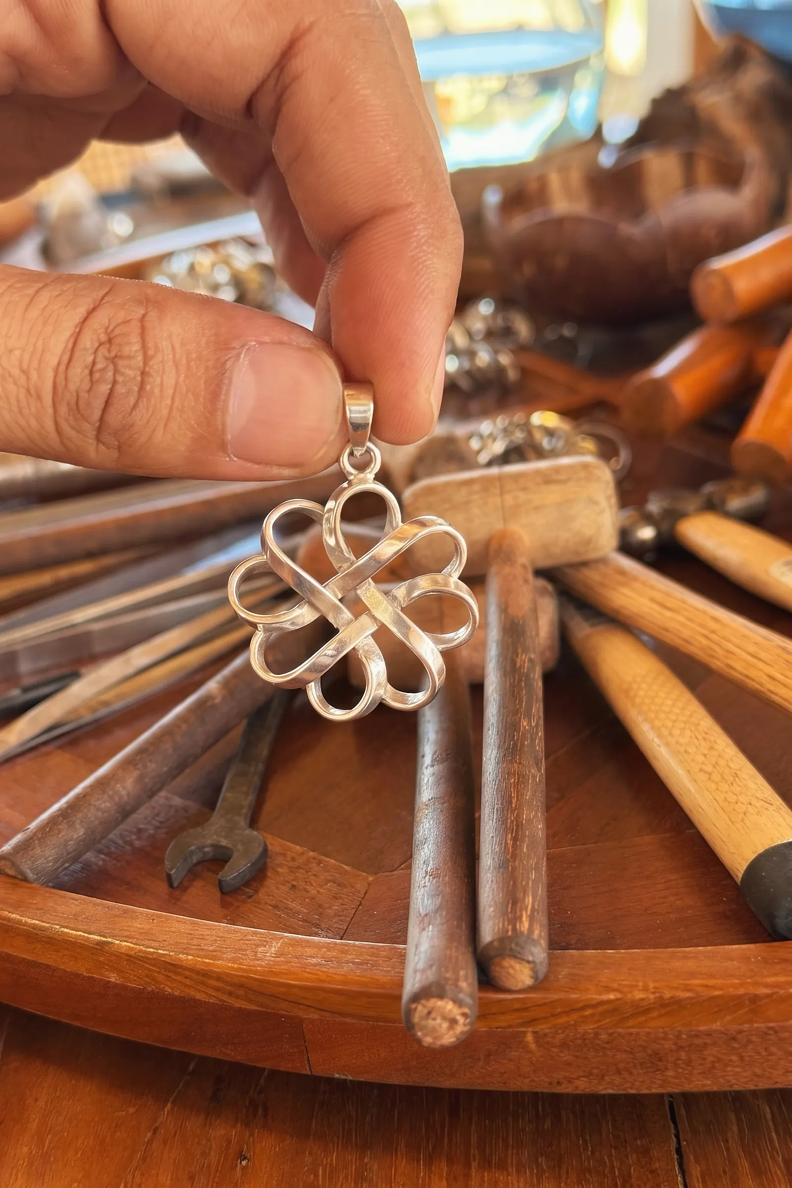 A silver knot pendant made in class, held up by its bail above the teak workbench